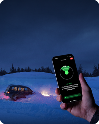 Photo d’une voiture bloquée dans la neige en pleine forêt pendant la nuit. À l’avant-plan, une main tient un iPhone 15. On peut voir à l’écran que la fonctionnalité Urgence SOS par satellite a établi une connexion